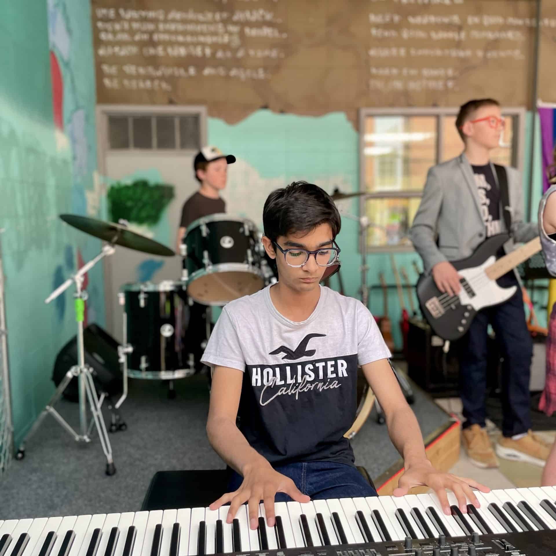 Ayush Shambhavi with Brokend Glasses Teenage boy with glasses and black hair plays keyboard with his rock band in the back. Performance is outdoors in a back alley.